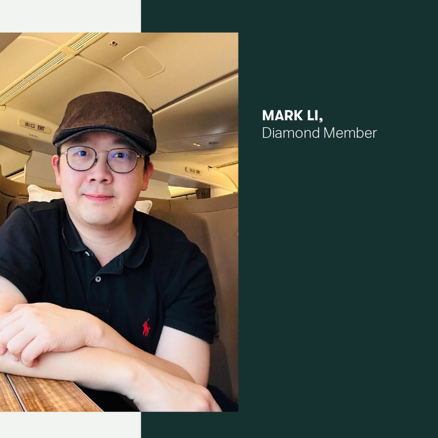 A man with glasses wearing a black polo shirt and a cap sits in the Business class cabin of a Cathay Pacific aircraft.